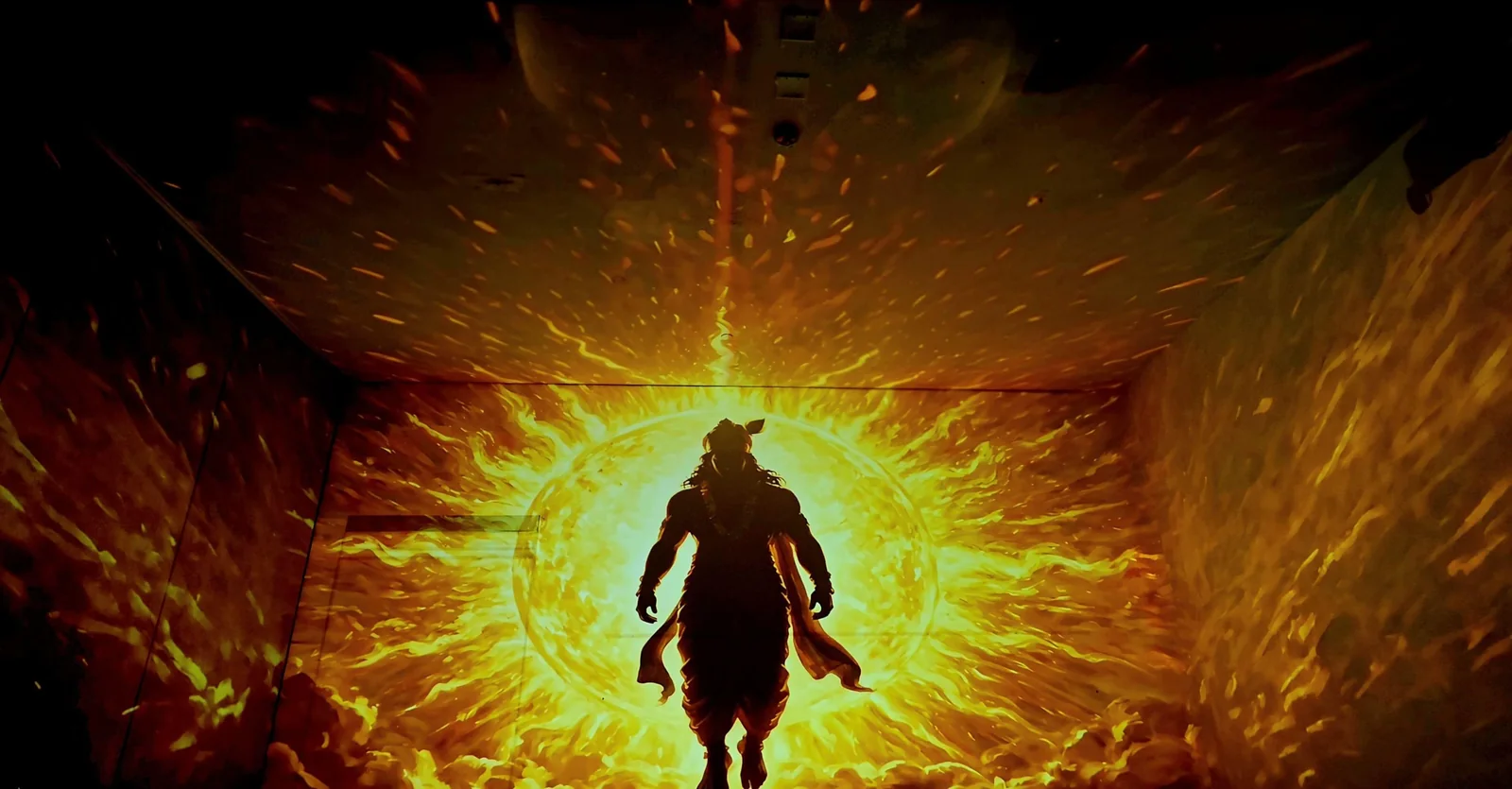 A room-scale projection: a silhouetted figure walking out of a blazing sun of fire, the light wrapping from the far wall onto the ceiling and side walls of the room